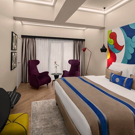 Sura Hagia Sophia Hotel Истанбул Екстериор снимка The photo shows a modern hotel room designed with a clean and vibrant aesthetic. It features a large bed with a decorative headboard and stylish bedding. Two purple armchairs are positioned beside a small table near large windows that allow natural l