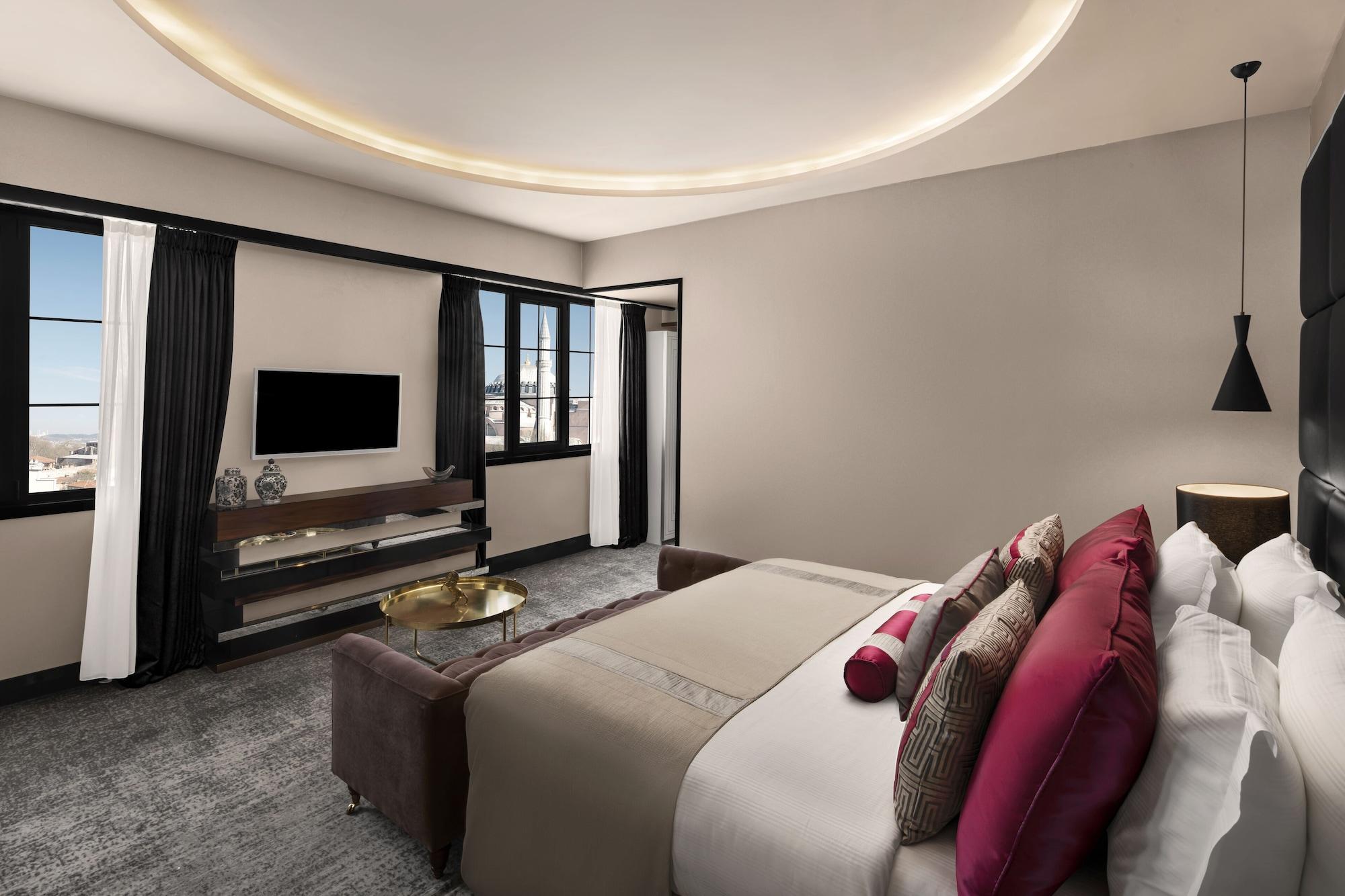 Sura Hagia Sophia Hotel Истанбул Екстериор снимка The photo shows a modern hotel room. It features a large bed adorned with decorative pillows in shades of pink and neutrals. There is a sleek wooden console beneath a flat-screen television mounted on the wall. The room has large windows with flowing