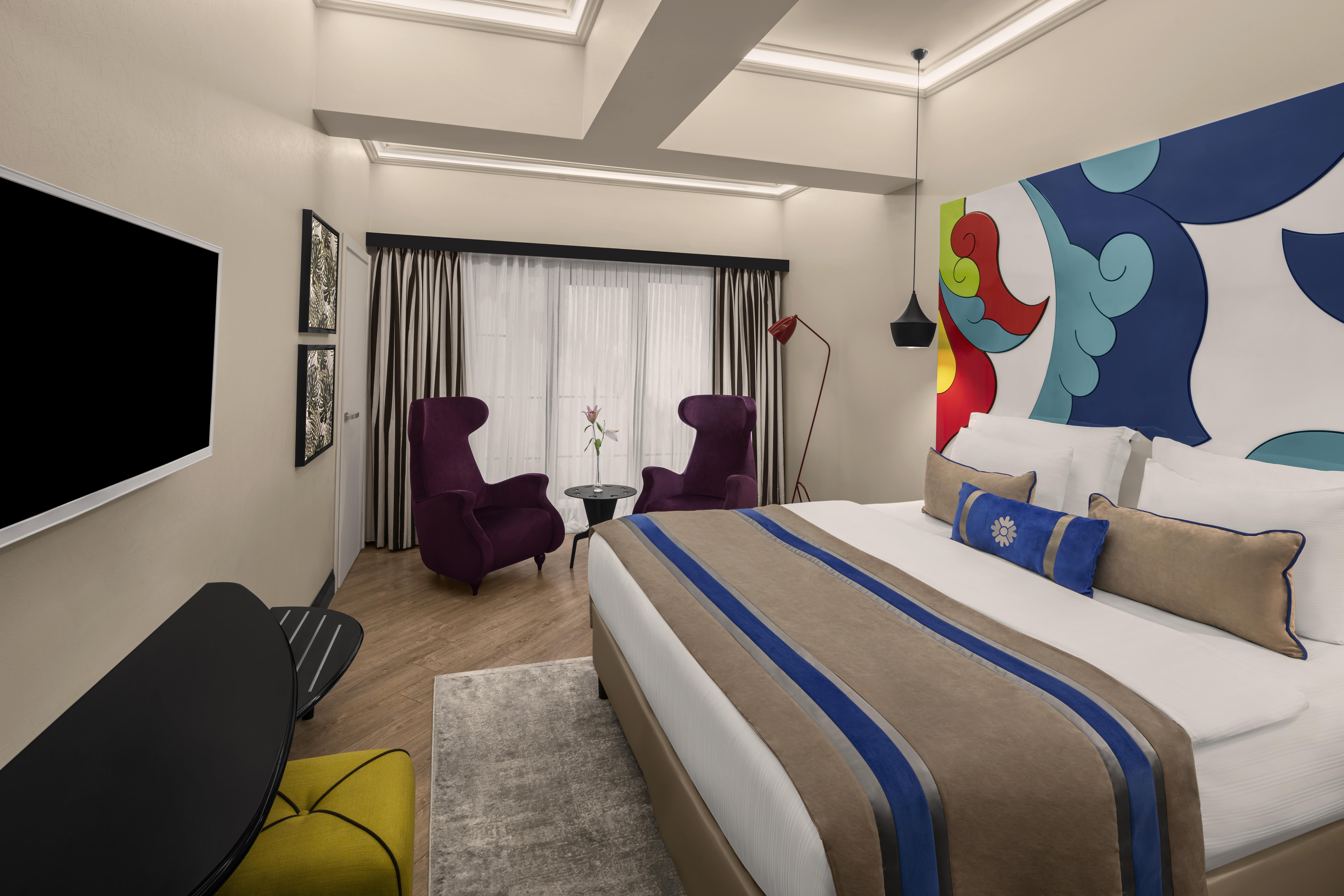 Sura Hagia Sophia Hotel Истанбул Екстериор снимка The photo shows a modern hotel room designed with a clean and vibrant aesthetic. It features a large bed with a decorative headboard and stylish bedding. Two purple armchairs are positioned beside a small table near large windows that allow natural l