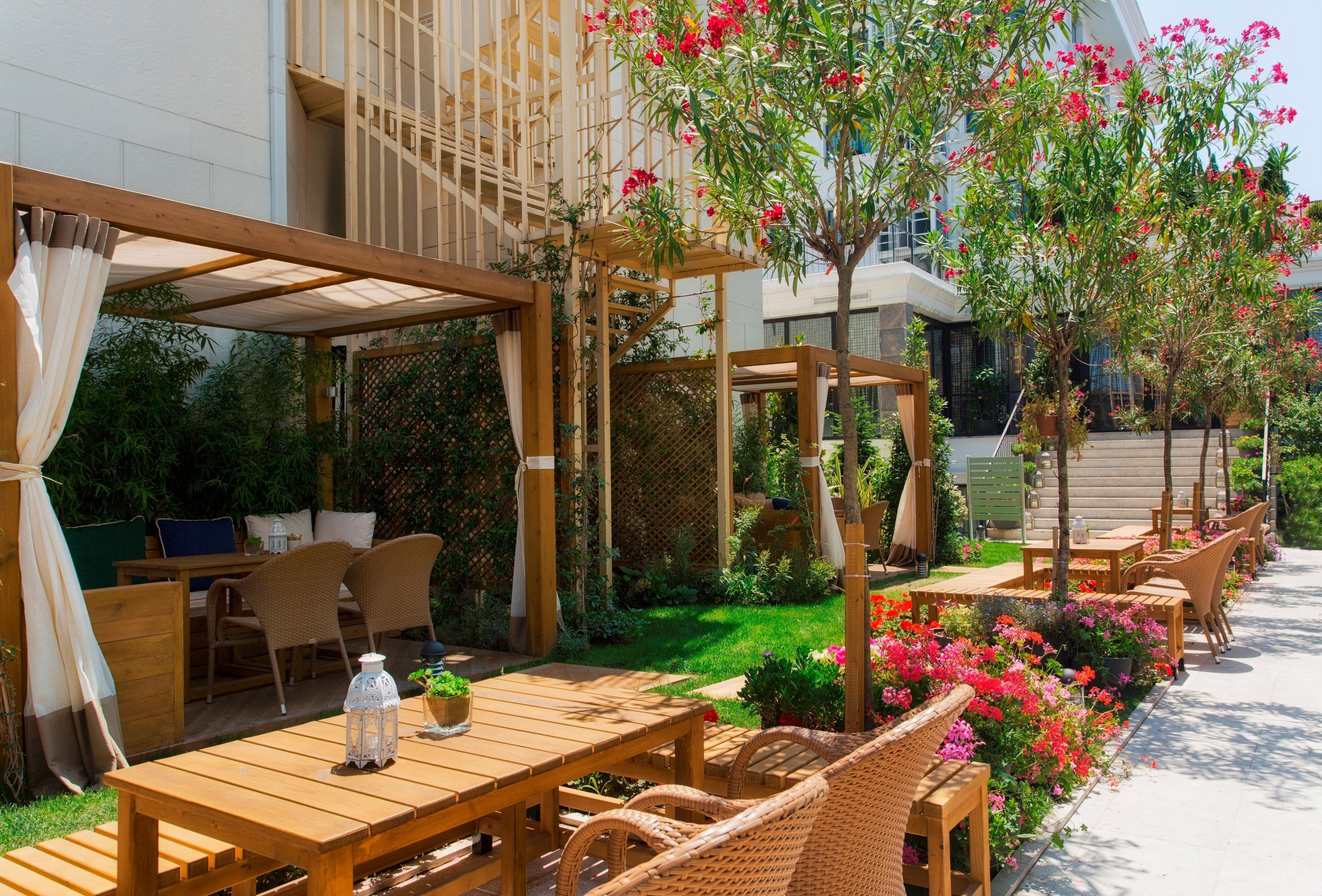 Sura Hagia Sophia Hotel Истанбул Екстериор снимка The photo shows a beautifully landscaped outdoor area with wooden furniture arranged for seating. There are tables and comfortable chairs made from woven materials. The space is decorated with lush greenery, including flowering plants and small trees