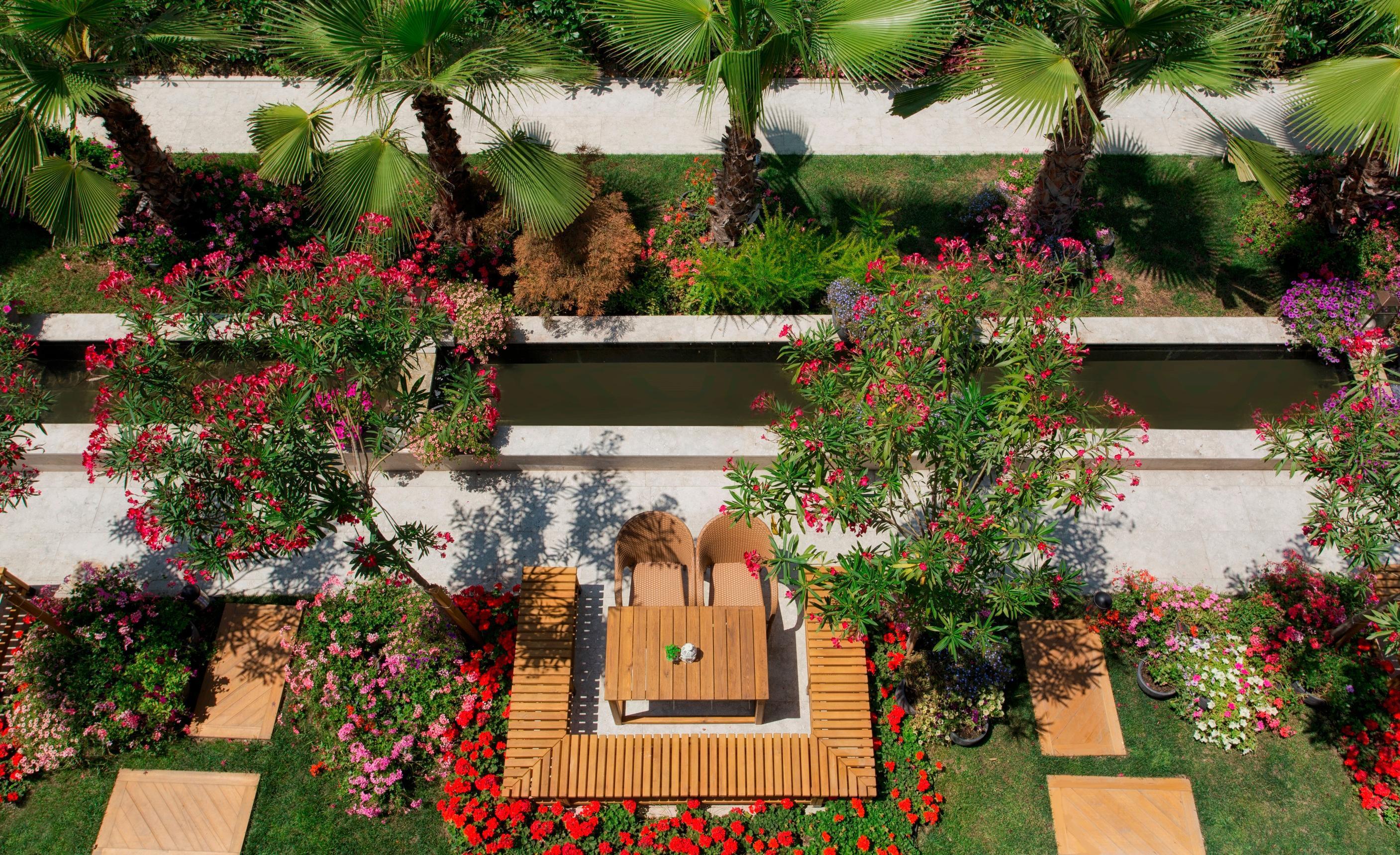 Sura Hagia Sophia Hotel Истанбул Екстериор снимка The photo depicts a beautifully landscaped garden area. In the center, there is a wooden table surrounded by chairs, situated on a deck that features a striped pattern. This seating area is framed by vibrant flowering plants and greenery, including p