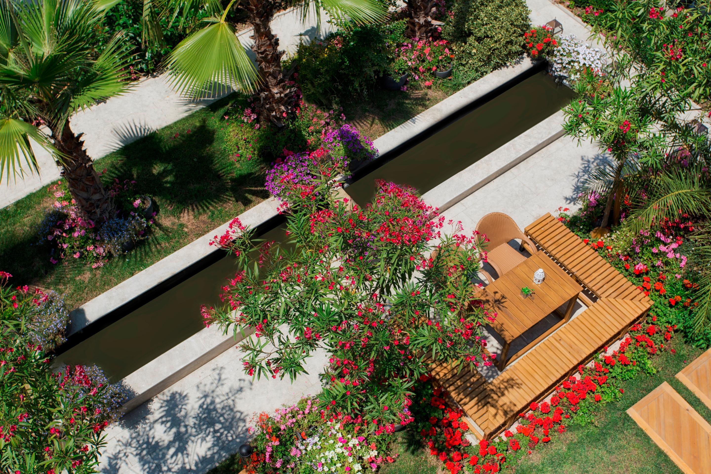 Sura Hagia Sophia Hotel Истанбул Екстериор снимка The photo shows a beautifully landscaped garden with vibrant flowers and greenery. There is a narrow, dark water channel running through the garden, flanked by lush palm trees and colorful flowering plants. A wooden deck or patio area, featuring a di