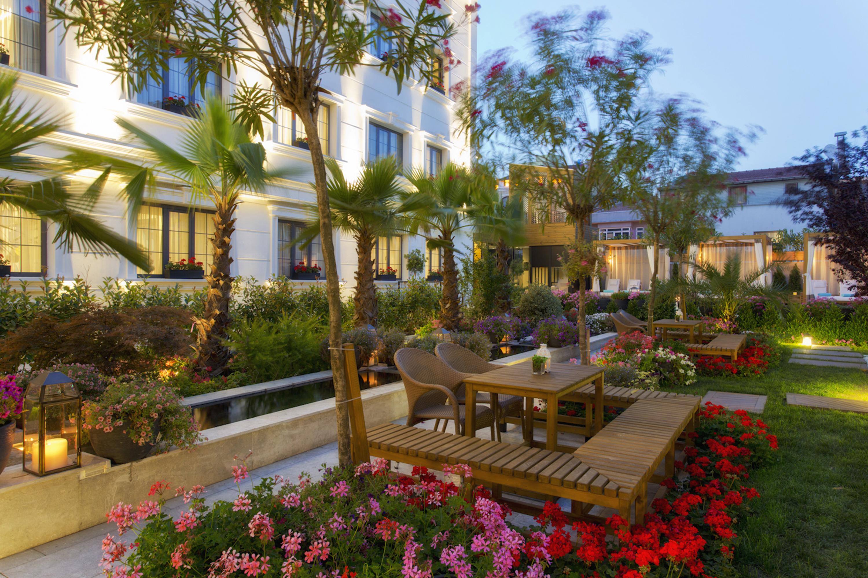 Sura Hagia Sophia Hotel Истанбул Екстериор снимка The photo depicts a beautifully landscaped garden area, likely at a hotel or a resort. It features wooden decking with seating arrangements, surrounded by vibrant flowers and greenery. There are palm trees and various plants, creating a tropical ambi
