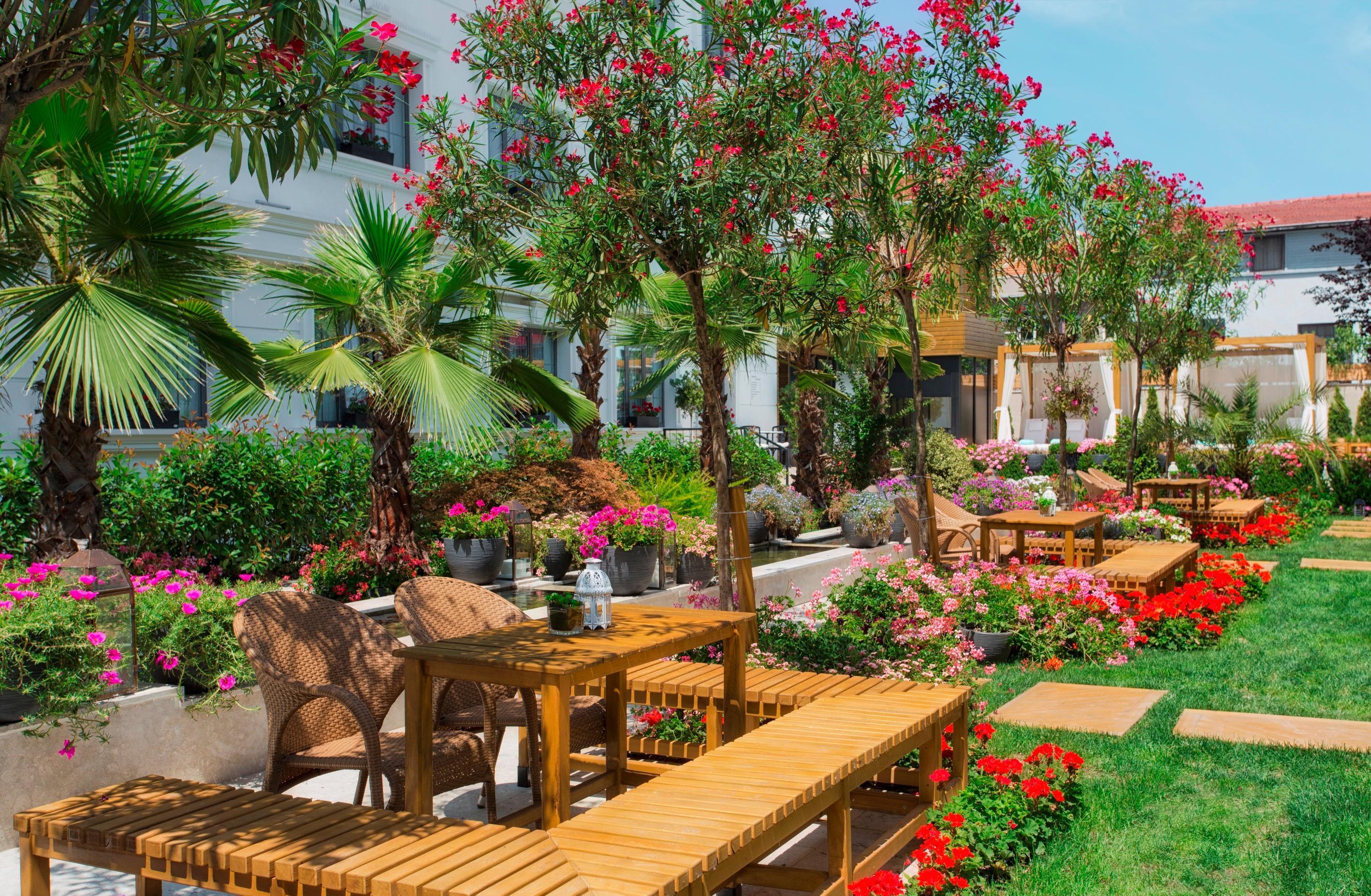 Sura Hagia Sophia Hotel Истанбул Екстериор снимка The photo depicts a beautifully landscaped outdoor garden area. There are several wooden benches and tables arranged among vibrant flowers and greenery. The garden features various types of plants, including palm trees and flowering bushes, creating 