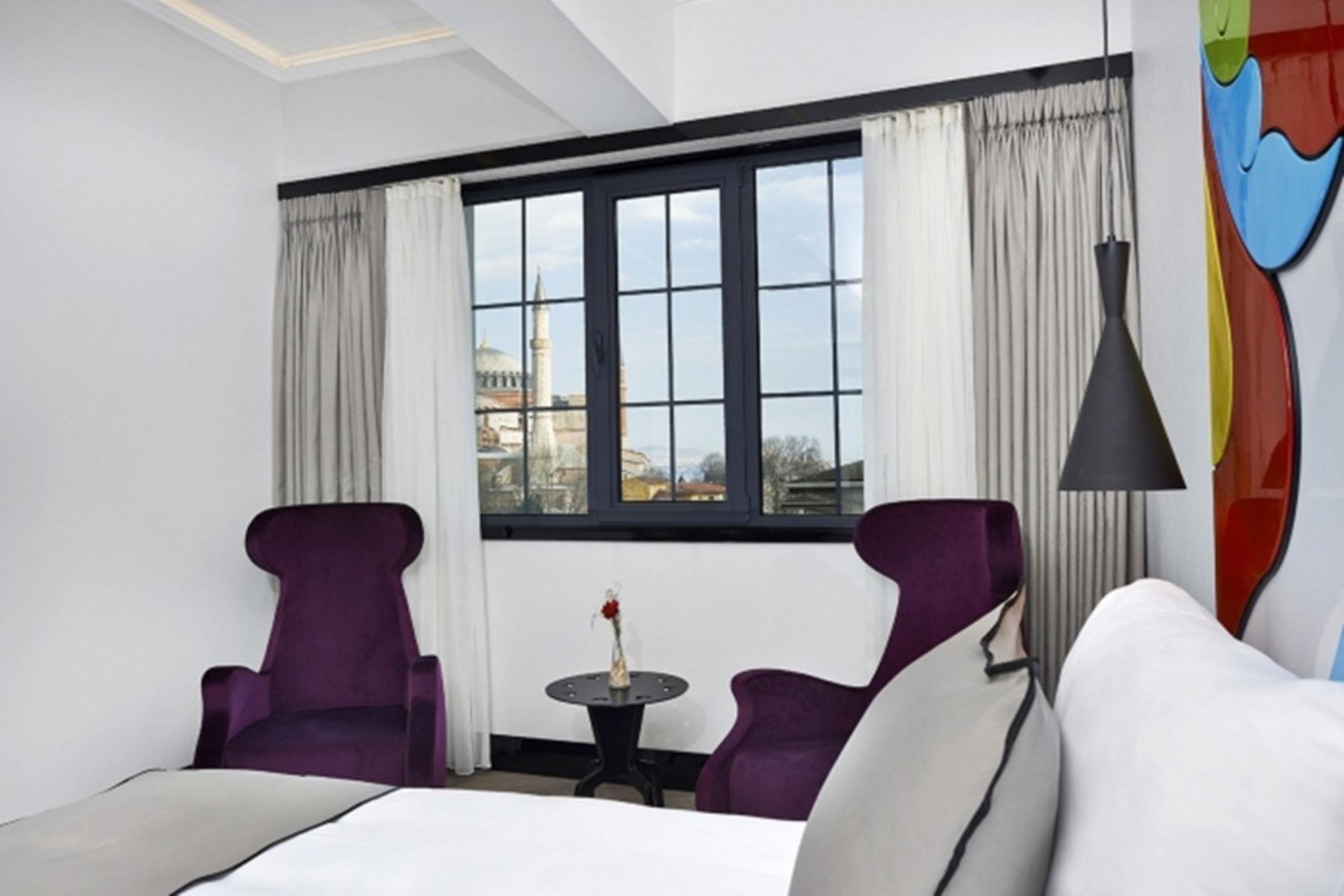 Sura Hagia Sophia Hotel Истанбул Екстериор снимка The photo showcases a modern hotel room with a stylish design. In the foreground, there are two purple armchairs placed near a small round table featuring a single flower. The room has large windows with white curtains, allowing natural light to floo