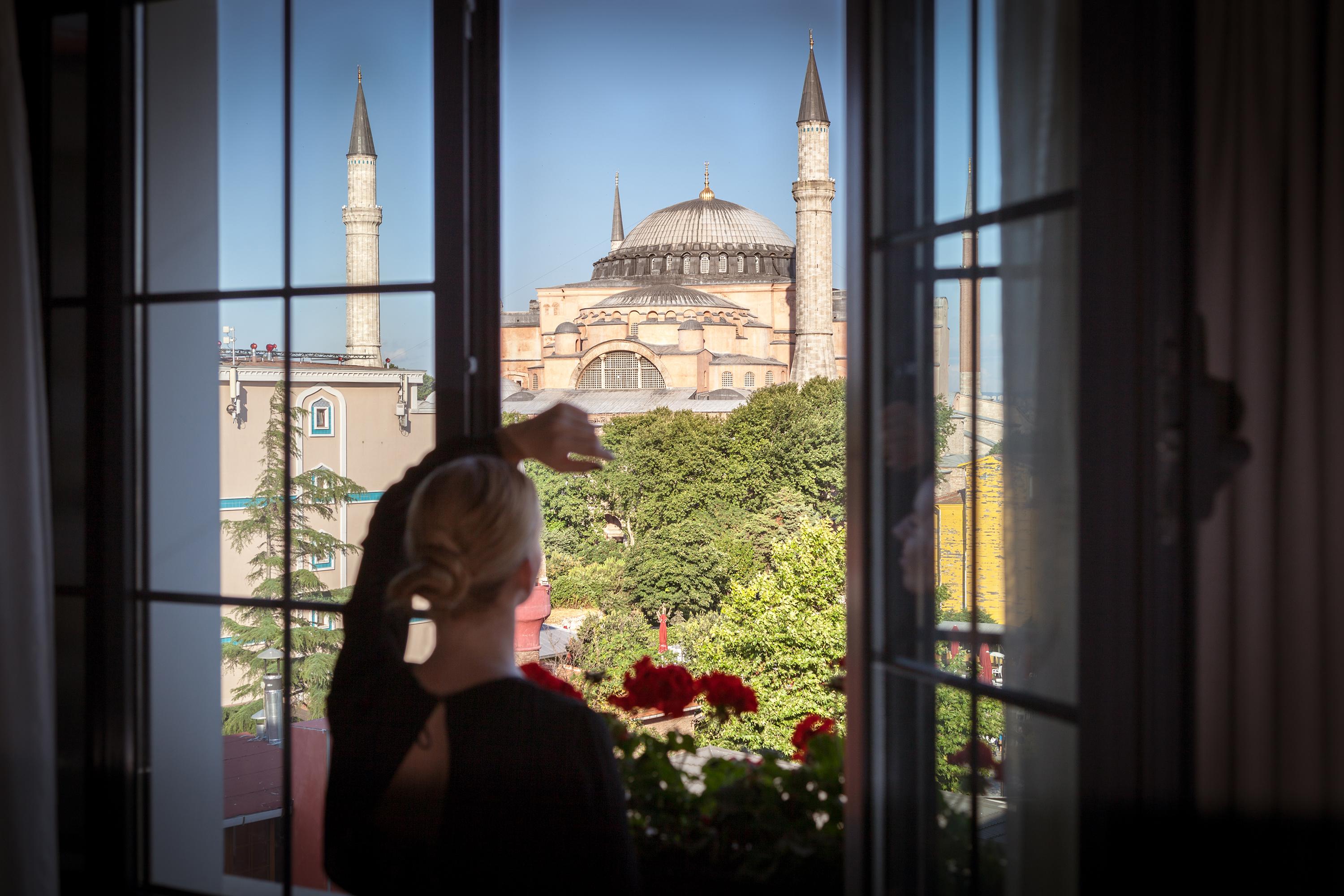 Sura Hagia Sophia Hotel Истанбул Екстериор снимка The photo shows a person standing by a window, gazing out at a view of a historic building, which appears to be the Hagia Sophia. This remarkable structure is characterized by its large dome and multiple minarets. The scene includes greenery and flow