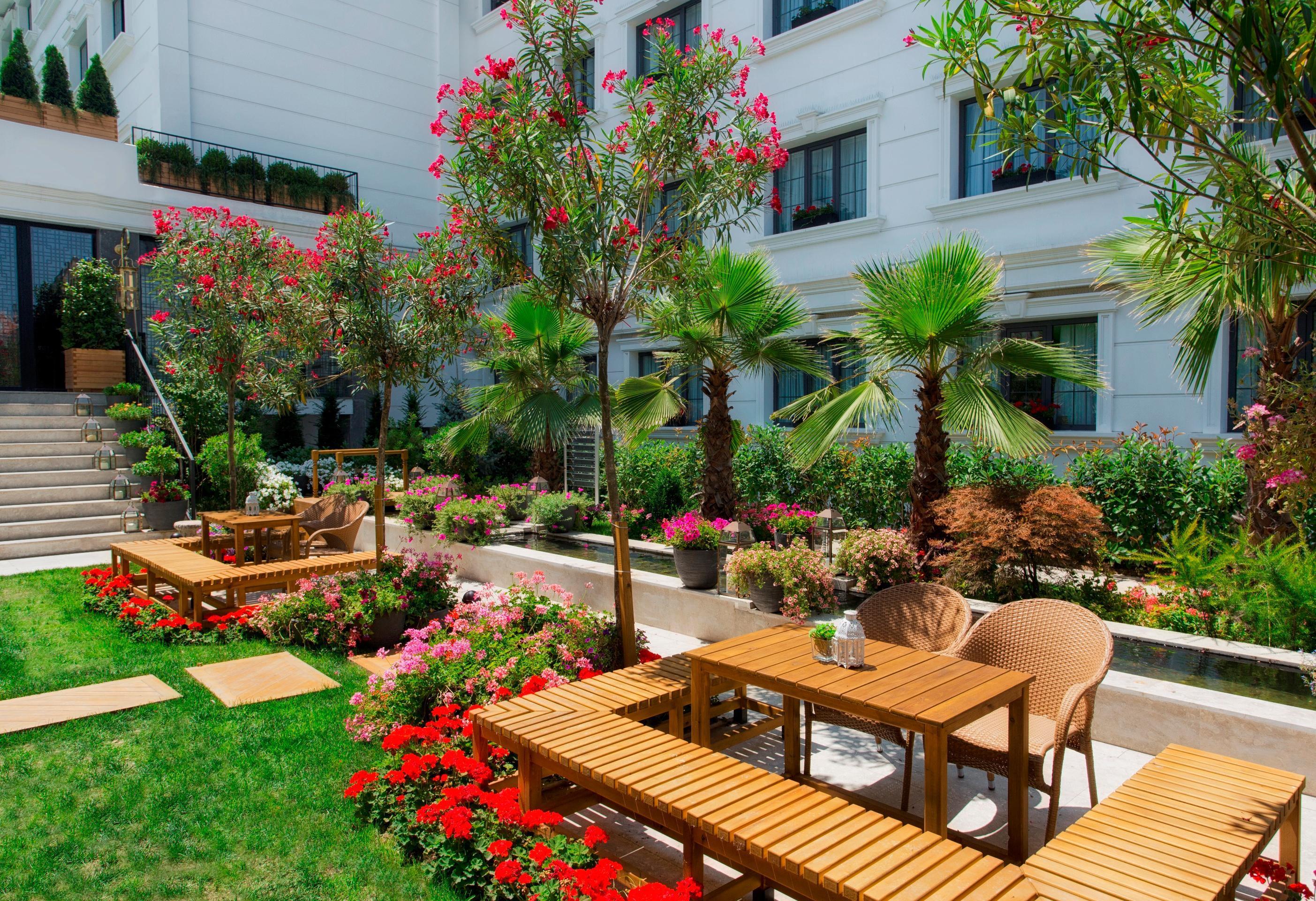 Sura Hagia Sophia Hotel Истанбул Екстериор снимка The photo depicts a beautiful outdoor garden area featuring well-maintained landscaping. It includes several wooden benches and tables arranged for dining or relaxation. Lush green grass contrasts with vibrant flower beds filled with colorful blooms.