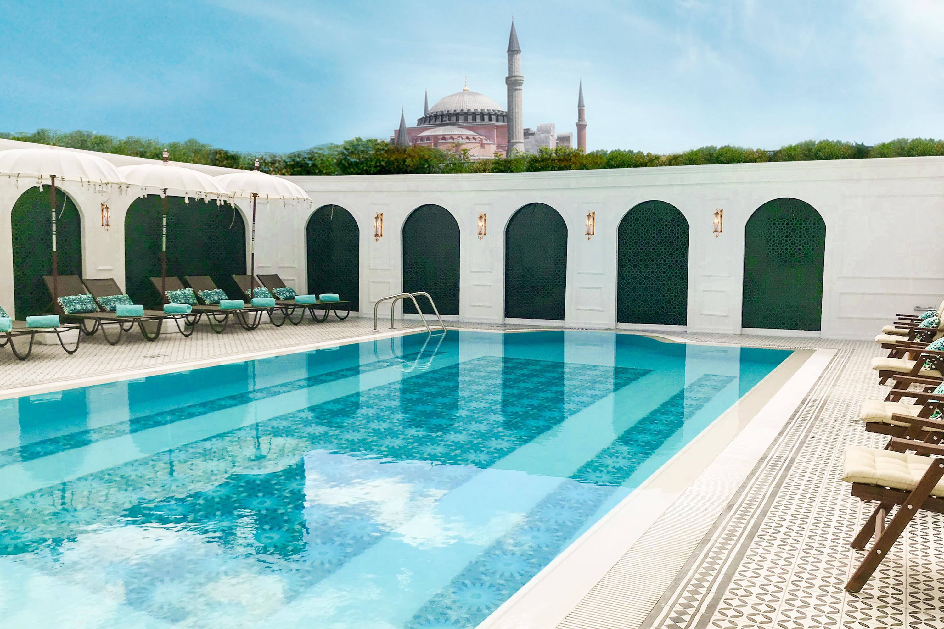 Sura Hagia Sophia Hotel Истанбул Екстериор снимка The image shows a swimming pool with clear, blue water. Surrounding the pool are several lounge chairs arranged neatly. In the background, there is an architectural landmark that resembles a historic mosque or cathedral, featuring tall minarets. The 