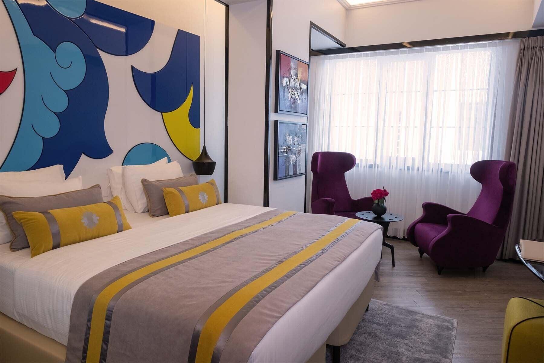 Sura Hagia Sophia Hotel Истанбул Екстериор снимка The photo shows a modern hotel room with a stylish design. It features a large bed with decorative pillows and a patterned blanket. The wall behind the bed has a colorful mural with blue and yellow shapes. Opposite the bed, there are two purple armch