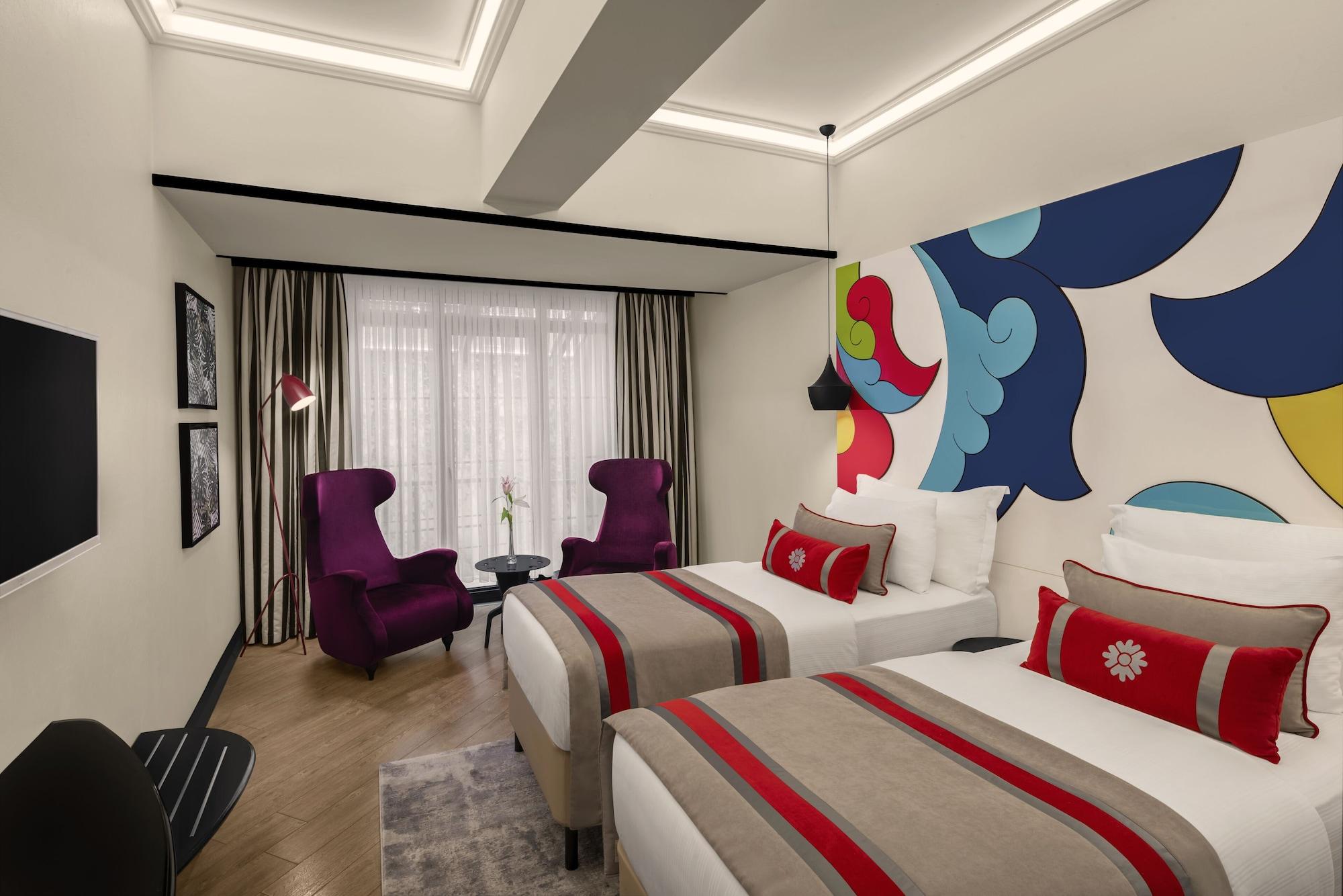 Sura Hagia Sophia Hotel Истанбул Екстериор снимка The photo shows a stylish hotel room featuring two beds with elegant white linens and decorative throw pillows. The room has modern decor, highlighted by a colorful mural on the wall. There are two purple armchairs placed near a small table, compleme