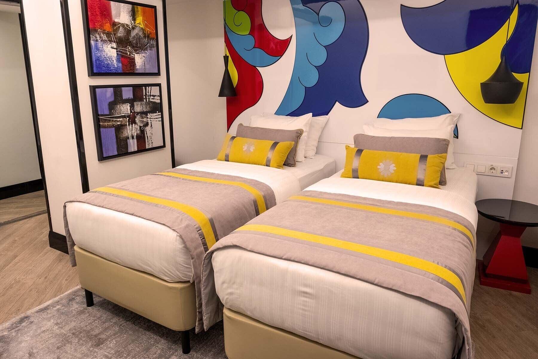 Sura Hagia Sophia Hotel Истанбул Екстериор снимка The photo shows a modern hotel room featuring two single beds with light-colored linens and decorative yellow-striped throw pillows. The headboards of the beds are against a colorful mural that includes abstract shapes in red, blue, and yellow. To th