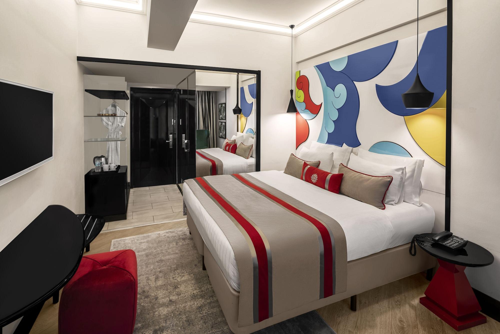 Sura Hagia Sophia Hotel Истанбул Екстериор снимка The photo shows a modern hotel room featuring a large bed with decorative pillows. The headboard has a bold, colorful mural depicting abstract shapes in blue, yellow, and red. There is a stylish black television mounted on the wall opposite the bed. 