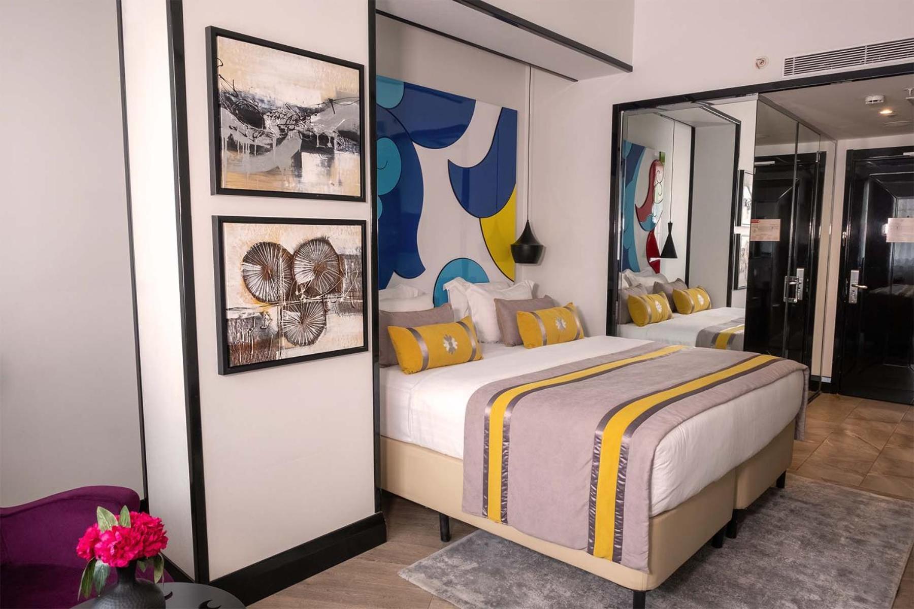 Sura Hagia Sophia Hotel Истанбул Стая снимка The photo shows a modern hotel room with a stylish design. There are two beds with a white duvet, complemented by colorful yellow and gray decorative pillows and a matching blanket. Behind the beds, there is a vibrant abstract mural featuring blue an